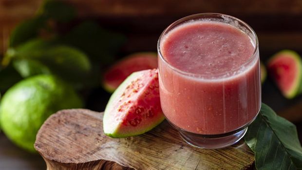 Delicious red guava juice alongside a slice of guava, in the background guavas and leaves on rustic wood