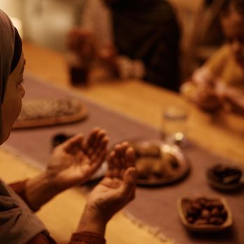 Close up of mature Muslim woman praying with her family at dining table.