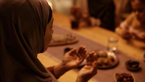Close up of mature Muslim woman praying with her family at dining table.