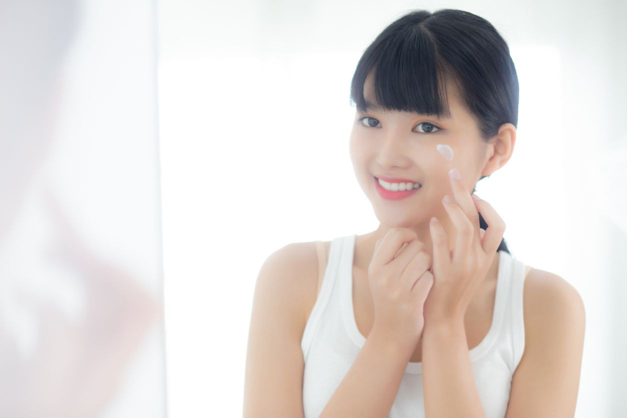 Beautiful young asian woman looking in mirror and apply moisturizer with cosmetic cream or lotion in bathroom at home, skincare for hygiene, treatment and bodycare, skin and anti-aging of facial.