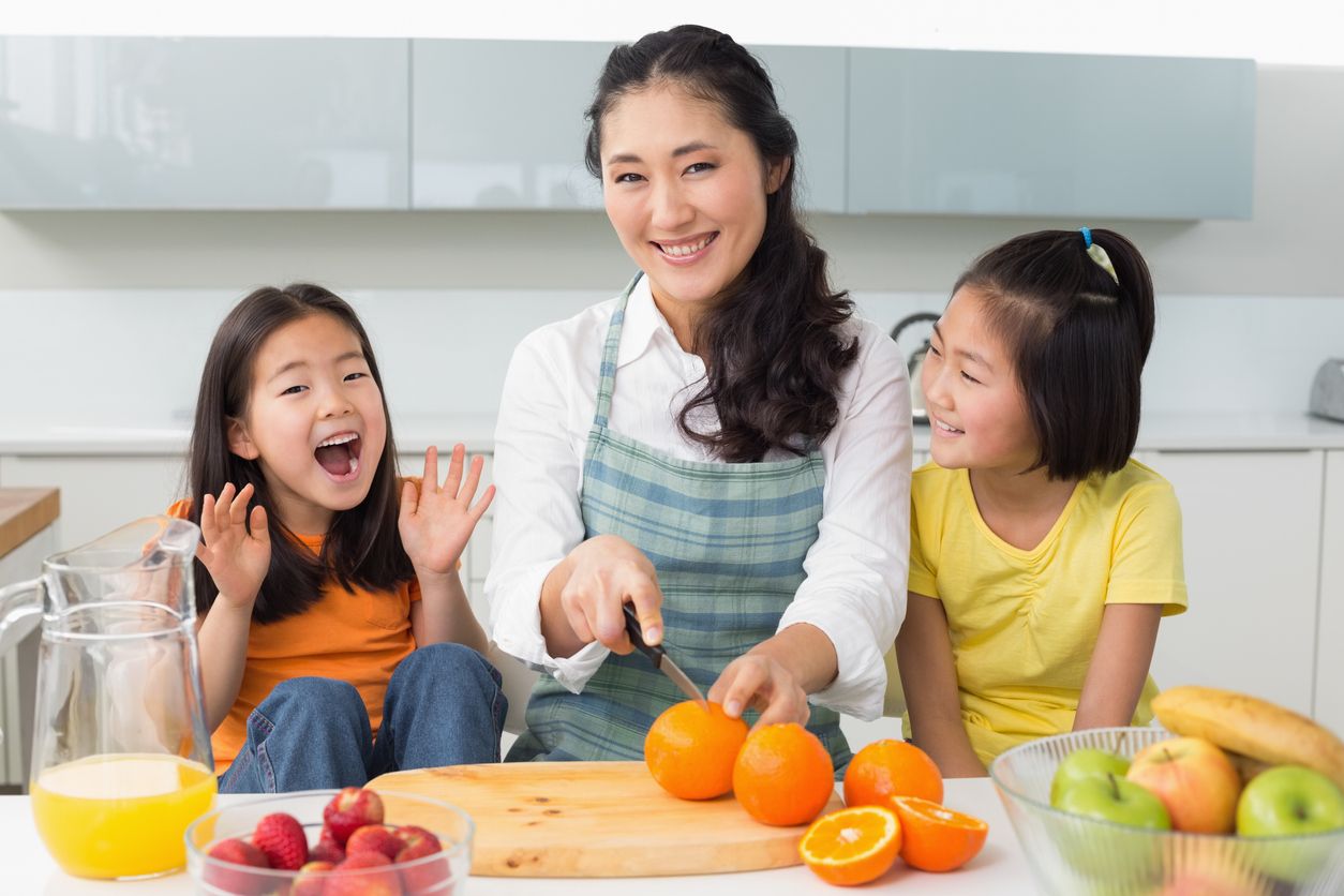 Portrait of a smiling woman with cheerful two daughters cutting fruit in the kitchen at home