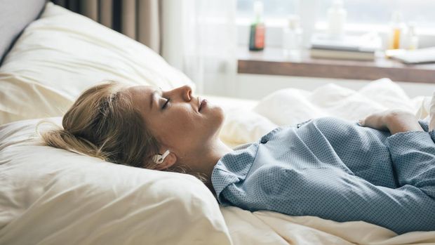 Happy blonde woman in blue pajamas lying in bed in the morning and listening to relaxing music.