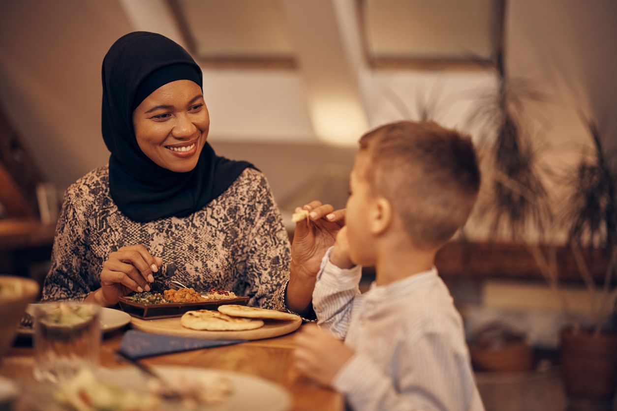 African American Muslim woman enjoying while feeding her son during a meal at dining table.