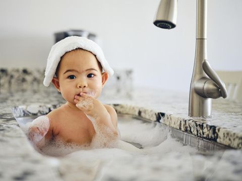 A 9 month old baby taking a bath in a kitchen sink.