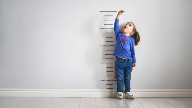 Little child is playing superhero. Kid is measuring the growth on the background of wall. Girl power concept.