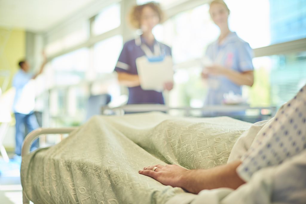 a female senior nurse or matron is  chatting to a young  a young student nurse is attending to the dressings trolley at the foot of a patient's bed . They are all defocussed apart from the patient's hand in the foreground.
