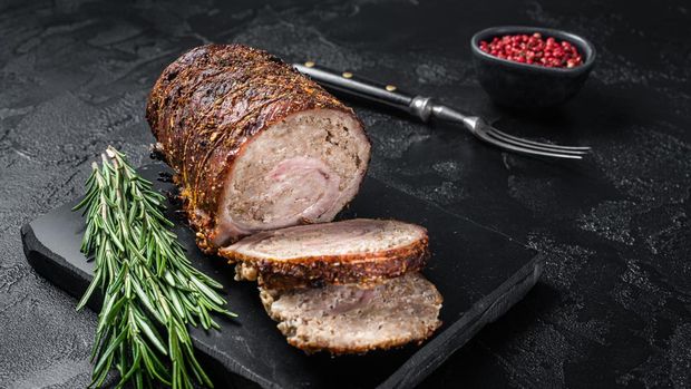 Baked pork roulade  Porchetta, rolled pork meat on a marble board with herbs. Black background. Top view.