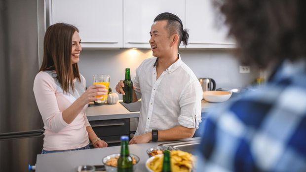 Over the shoulder view of young Asian male and Caucasian female enjoying drinks and conversation in kitchen at social gathering.