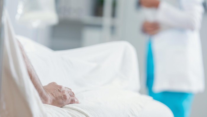 Ilustrasi dirawat Unrecognizable senior mans hand rests on hospital bed while waiting on his test results. A doctor is blurred in the background.