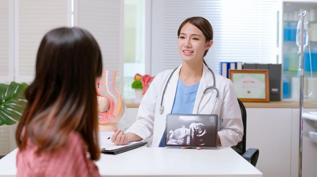 A pregnant female of Asian decent, lays out on an exam table as a technician conducts her ultrasound.  She is dressed casually and has her belly exposed as she looks to the screen to see her baby.