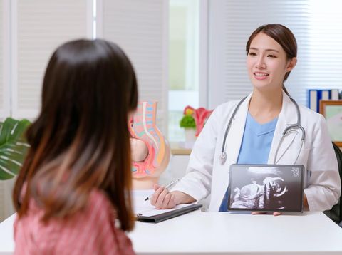 A pregnant female of Asian decent, lays out on an exam table as a technician conducts her ultrasound.  She is dressed casually and has her belly exposed as she looks to the screen to see her baby.