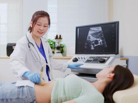 A pregnant female of Asian decent, lays out on an exam table as a technician conducts her ultrasound.  She is dressed casually and has her belly exposed as she looks to the screen to see her baby.