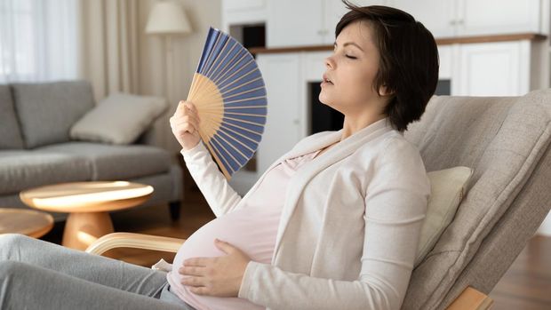 Young latin woman pregnant using hand fan sitting on sofa at home