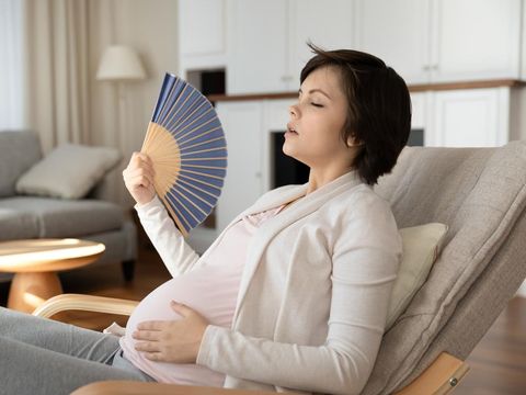 Young latin woman pregnant using hand fan sitting on sofa at home