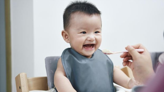 Happy infant Asian baby boy eating food by himself on baby high chair and making mess with copy space.