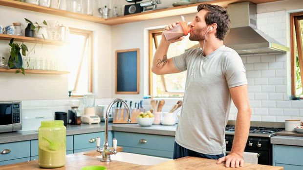 One fit young caucasian man drinking bottle of chocolate whey protein shake for energy for training workout while wearing earphones in a kitchen at home. Guy having sports supplement for muscle gain and dieting with weightloss meal replacement