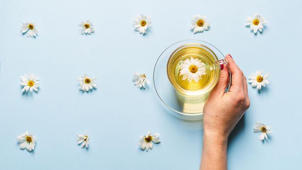 A mug of flowered chamomile tea in a woman's hand on a blue background with flowers spread out. Floral summer background. The concept of the arrival of summer mood and health. Flat lay.