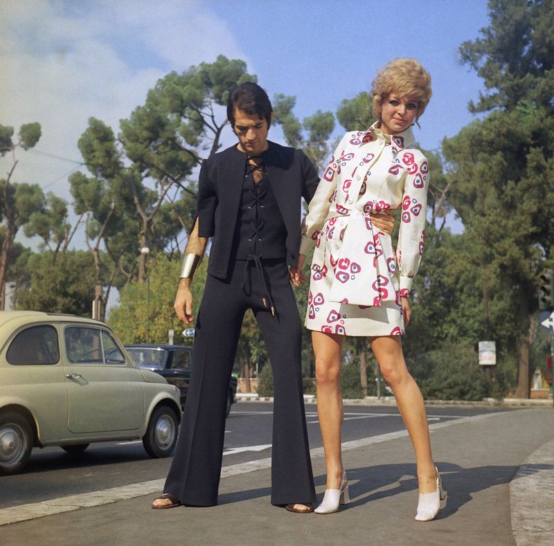 Two creations for the beach by the Angelo Litrico fashion house of Rome, Nov.  3, 1969, to be presented at the Florence show of Italian spring-summer 1970 high fashion ready-to-wear boutique collections, opening on November 3. For the man: An evening dress fir the beach of blue jersey, consisting of a sort of overalls with very wide legged trousers, laced along the front and worn with a small jacket and a large gilded bracelet.  For the Woman: An after the sea beach dress of silk shantung printed on ivory ground, with pockets and a scarf tied around the waist.  (AP Photo/Mario Torrisi)