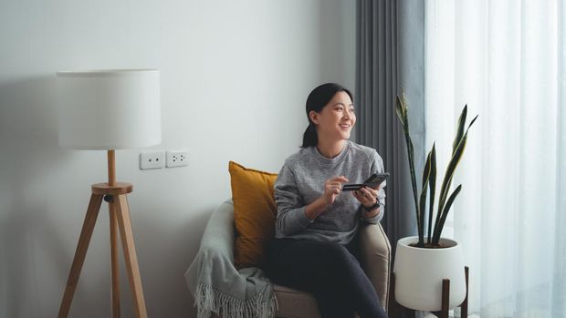 Asian woman sitting on armchair in living room using smart phone and credit card for shopping online at home. Happy woman shopper using instant mobile payments making purchase in online store.