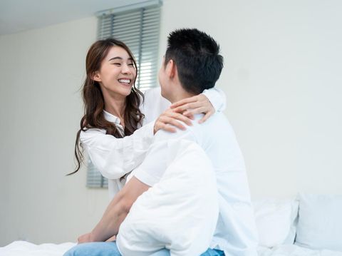 Asian new marriage couple sit on bed and look at each other with love. Attractive beautiful young man and woman in pajamas enjoy early morning activity in bedroom at home. Family relationship concept.