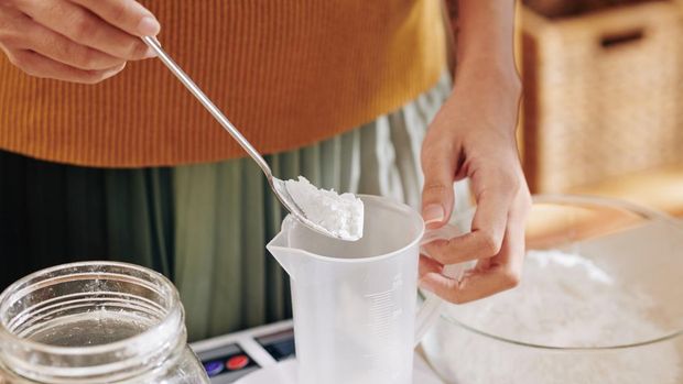Close-up image of woman putting spoon of lye in plastic jar when making soap at home