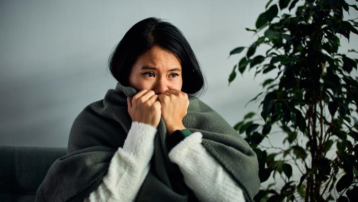 ilustrasi dingin A sad and ill Asian female wrapped herself in a warm grey blanket, sitting on a couch at home.