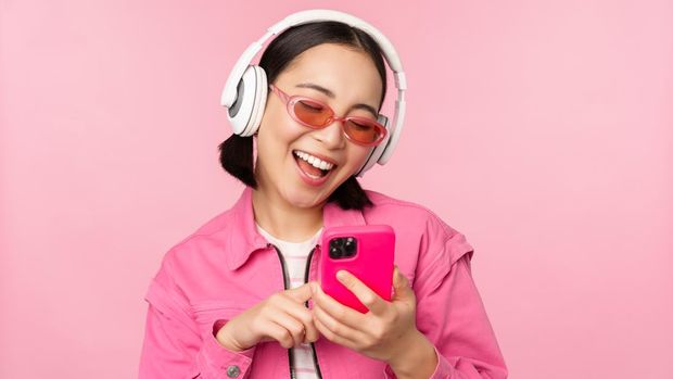 Stylish asian girl dancing with smartphone, listening music in headphones on mobile phone app, smiling and laughing, posing against pink background.