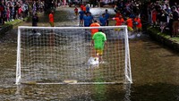 Dua tim yang beranggotakan enam orang memainkan pertandingan sepak bola di air sungai yang dangkal.