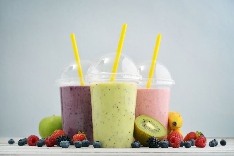 Fruit smoothies in plastic cups with blueberry, strawberry, kiwi, blackberry, raspberry and banana on blue background. Take away drinks concept.