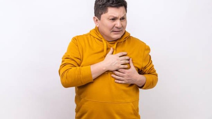 Ilustrasi nyeri dada sebelah kanan. Heart attack or problem. Portrait of sad shocked handsome man standing and holding his chest with painful face, wearing urban style hoodie. Indoor studio shot isolated on white background.