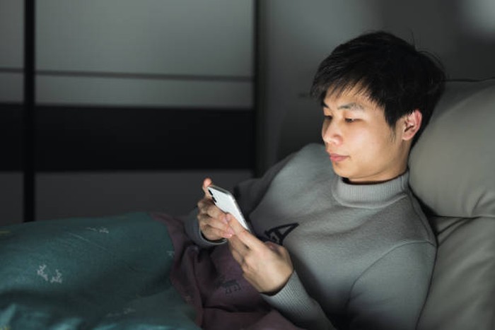 At night, a young Asian man sits on his bed and plays with his mobile phone, the screen light illuminating his face.