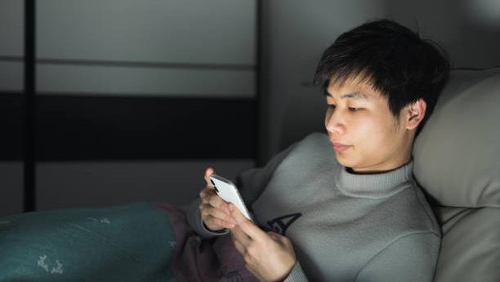 Ilustrasi begadang sambil main HP At night, a young Asian man sits on his bed and plays with his mobile phone, the screen light illuminating his face.