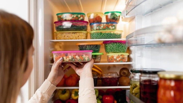 Close up shot of woman taking container with frozen mixed vegetables from refrigerator while looking at camera. Penyebab Kulkas Tak Dingin