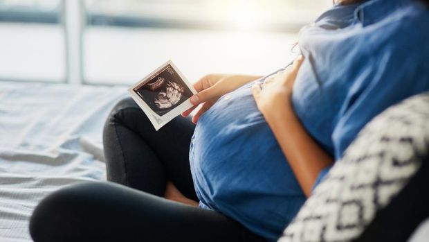 A photo with the focus on the unrecognizable pregnant woman in the foreground as the unrecognizable doctor shows her an ultrasound on a digital tablet in the background.