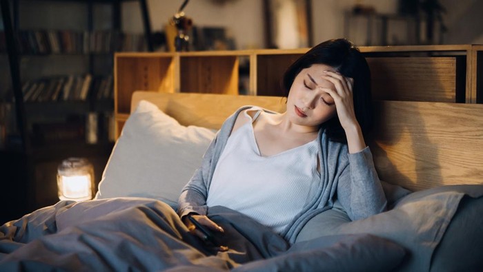 Ilustrasi tak bisa tidur atau sulit tidur Young Asian woman feeling sick and suffering from a headache, lying on the bed and taking a rest at home