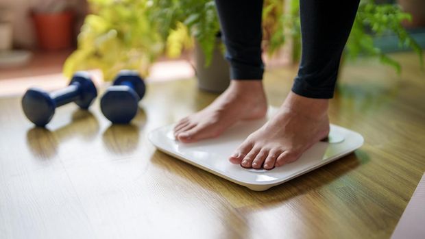 A close-up view of a young woman standing on a weighing scale, focusing on her feet and the display as she checks her weight. The scene highlights the importance of health and self-awareness in maintaining a balanced lifestyle.
