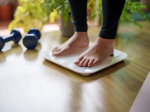 A close-up view of a young woman standing on a weighing scale, focusing on her feet and the display as she checks her weight. The scene highlights the importance of health and self-awareness in maintaining a balanced lifestyle.