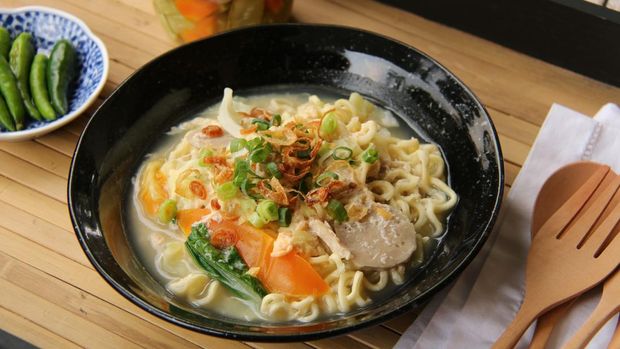 Mie Godog Jawa, the popular Javanese-Peranakan street food of noodle soup, served fancily on ceramic soup plate which then placed on a contemporary bamboo tray. The dish is also served with cucumber pickles and green chili peppers. Pairs of wooden cutlery are set on a white embroidered napkin which is placed next to the plate.