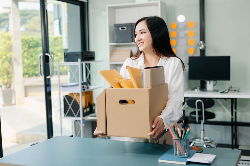 Happy and excited young beautiful Asian woman office worker celebrating her resignation, carrying her personal stuff. leaving job, changing or company.in modern office
