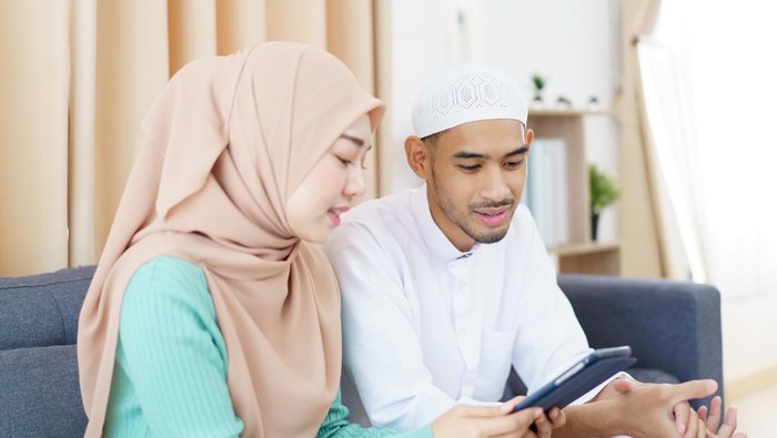 Asian Muslim couple is sitting together in the living room, reading and studying the Alquran during the fasting month of Ramadan. Asian muslim man and wo suami istri bersinggungan man are reading the Koran on a digital tablet.