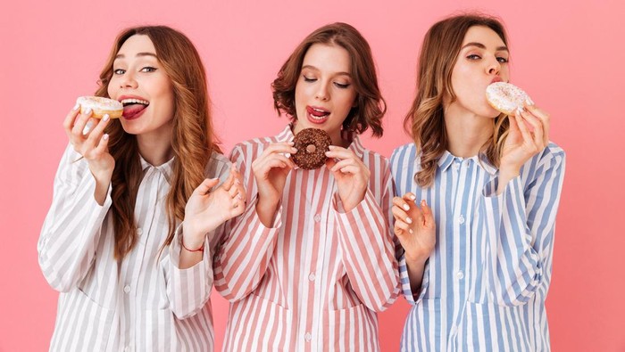 Picture of amazing three women friends wearing leisure clothings eating donuts and having fun at pajamas party isolated over pink background