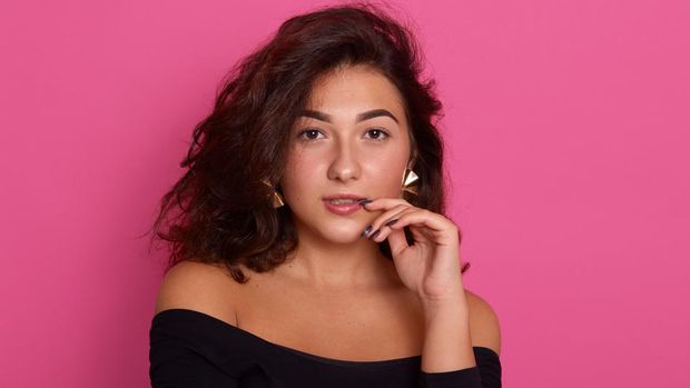 Thoughtful female keeps fore finger near lips, being deep in thoughts, dresses black shirt isolated over pink background with blank copy space for your advertisement, lady with confident expression.
