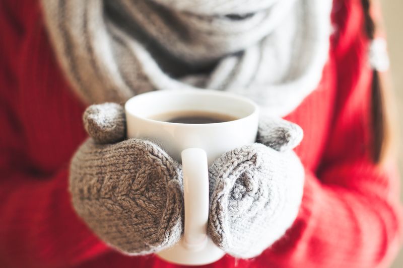 505578044 Female hands holding hot mug close up. Woman hands in woolen mittens holding a cup with hot cocoa, tea or coffee.