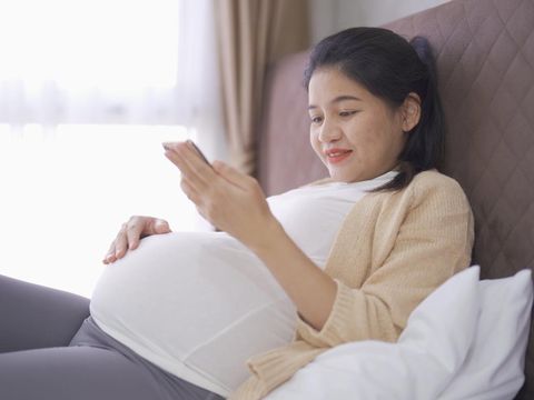 A portrait of happy Asian pregnant woman using online mobile phone technology device with screen in bedroom at home. Having a baby. Family people lifestyle. Mom love.