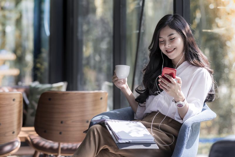 Shot of a beautiful young asian woman holding cup of coffee and listening music from her phone in the coffee shop