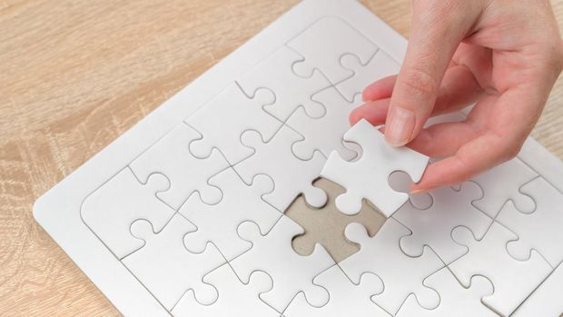 Female hand putting a missing piece and solving blank white jigsaw puzzle placed on top of old wooden oak table, close up, selective focus