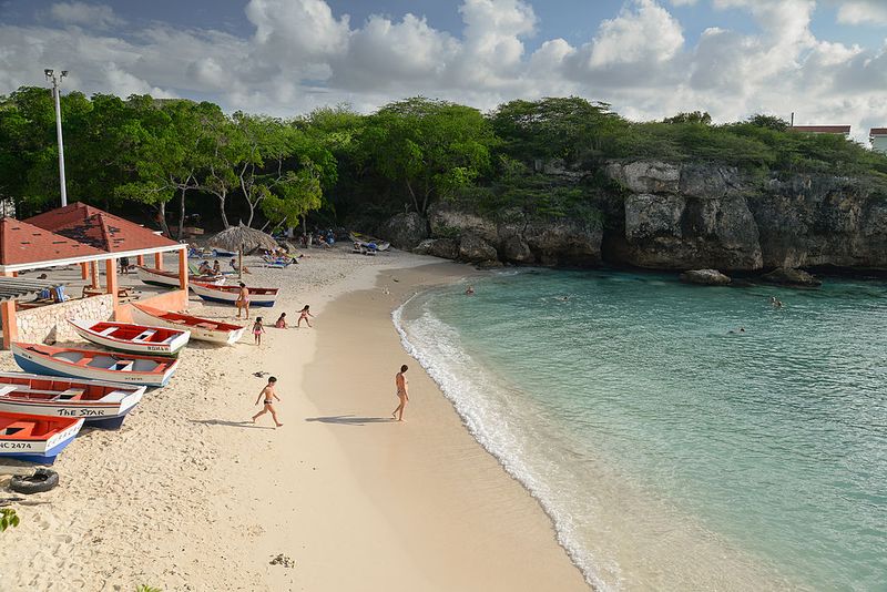 [UNVERIFIED CONTENT] View from the top of the cliffs at Lagun Beach towards the  beach, Curacao, Netherland Antilles.A couple of persons can be seen walking towards the Caribbean's pristine and turquoise water. Also, it can be seen some colorful fisherman boats on the left side of picture.The rocky cliffs and a partially cloudy sky can be seen at the background.