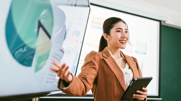 A professional Asian business woman presents data on screens in a board meeting room, engaging her audience with confidence and clarity. A businessman wearing a blazer while using a tablet for meeting