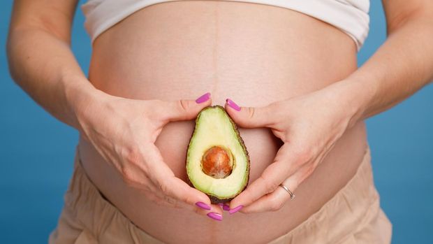 1205487033 Studio shot of young pregnant woman holding one half of a fresh avocado fruit close to her belly against blue background - healthy nutrition and pregnancy concept