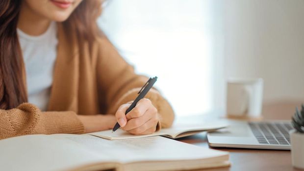 Close up - Hand of woman writing in spiral notepad placed on wooden desktop with various items at home. Copy space